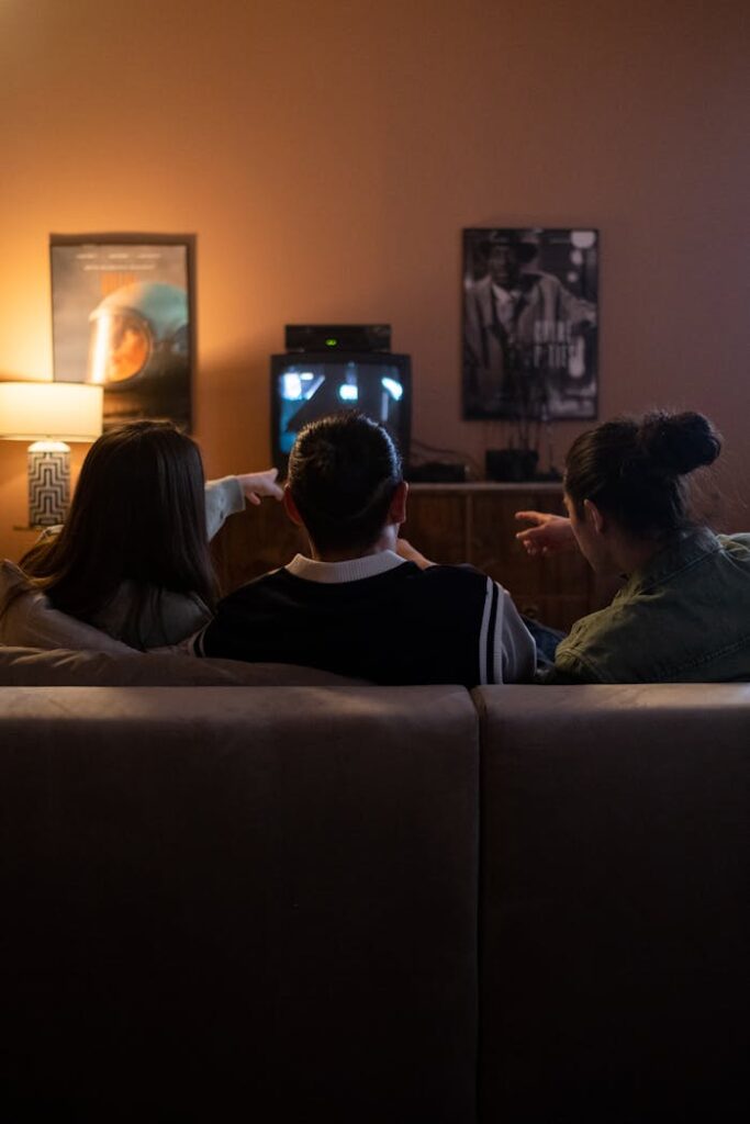 Three friends sitting on a couch enjoy a cozy movie night together, pointing at the TV screen.